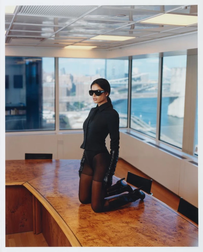Model kneeling on a wooden boardroom table wearing a black button-down shirt, tights, sunglasses, and gloves.
