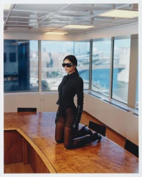 Model kneeling on a wooden boardroom table wearing a black button-down shirt, tights, sunglasses, and gloves.