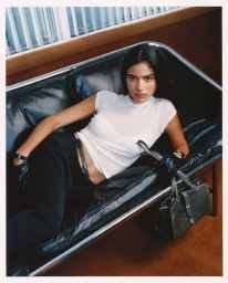 Model lying on a black leather sofa wearing a white t-shirt, dark trousers, gloves, and holding a dark handbag.