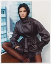 Model Rania Benchegra wearing a dark leather jacket and gloves seated against a large office window.