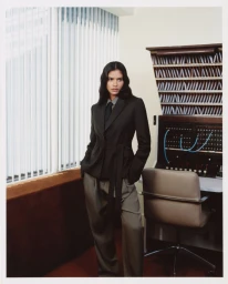 Model Rania Benchegra in a dark belted blazer and trousers standing in an office with vertical blinds for Kith Women.