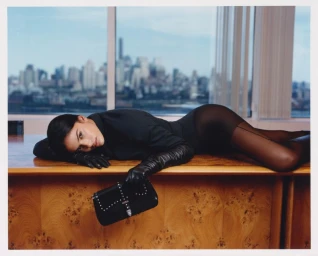 Model lying on a wooden office desk wearing black clothing, leather gloves, and holding a studded handbag with a city skyline view behind.
