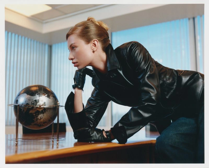 Model wearing a black leather jacket and gloves leaning over a wooden desk with a globe in an office setting.