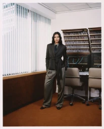 Model wearing a dark blazer and grey trousers standing in an office interior with vertical blinds and a switchboard.