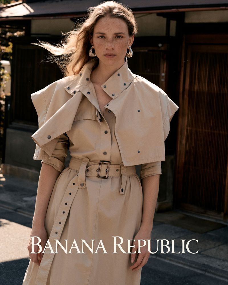 Model in a tan belted trench coat and silver earrings standing outdoors for Banana Republic.