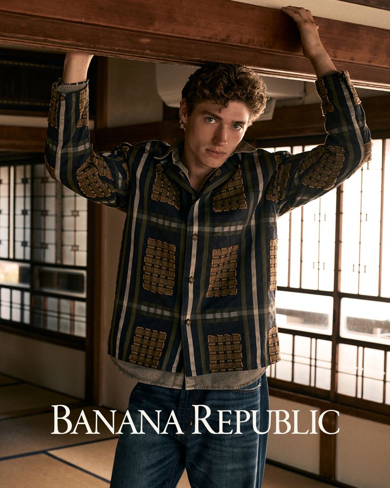 Valentin Humbroich wearing a plaid shirt and blue jeans, holding onto overhead wooden beams inside a traditional interior.