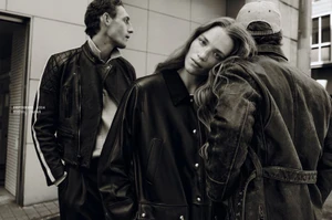 Monochrome fashion campaign image featuring three models wearing dark leather jackets against an urban building exterior background.