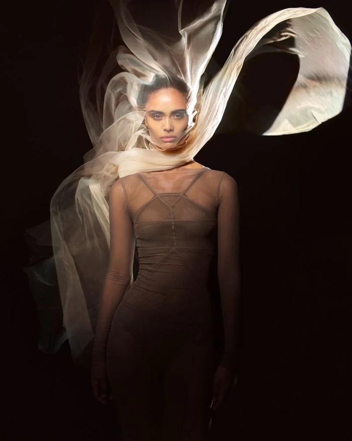 Model in a fitted brown sheer garment with a billowing translucent cream fabric framing her face against a black background