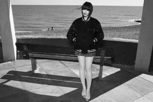Black and white portrait of a woman standing by a park bench overlooking the sea, wearing a varsity jacket