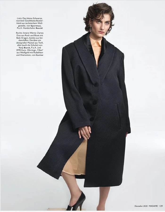 Model wearing a long black coat over a peach dress and black heels against a plain white background