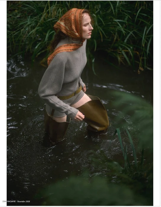 Model wearing a patterned headscarf, grey top, and tall waders standing in a river surrounded by lush green riverbank vegetation.