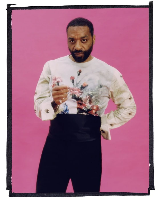 Chiwetel Ejiofor wearing a floral print top and high-waisted black trousers against a pink background