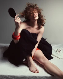Woman holding a small hand mirror with red bangles on her wrist sitting on a white sheet against a grey background