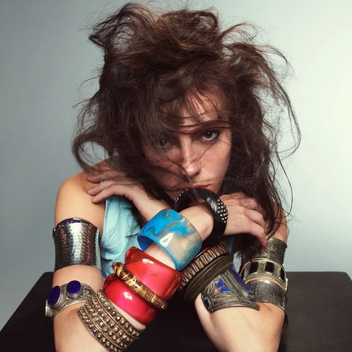 Close-up portrait of a woman with tousled dark hair wearing multiple chunky bangles and cuffs on both arms against a plain backdrop.
