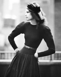 Black and white portrait of a female model wearing a dark long-sleeve dress and a distinctive cap outdoors.