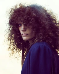 Close-up portrait of a model with voluminous curly brown hair and a blue garment against a bright background.