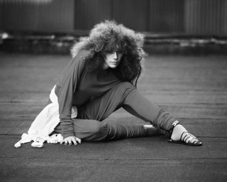 Model with voluminous curly hair seated outdoors wearing long-sleeved top, leggings, and strappy sandals for System Issue No. 26
