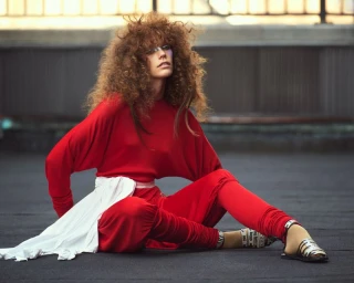 Model with voluminous curly hair wearing a vibrant red outfit seated outdoors on a dark rooftop