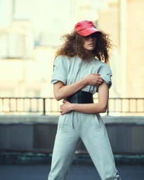 Model in grey jumpsuit with a black wide belt and red cap standing outdoors on a rooftop with a railing.