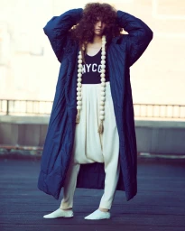 Model with voluminous curly hair wearing a long navy quilted coat, white drop-crotch trousers, and a black tank top outdoors.