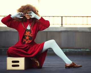 Model with voluminous curly hair wearing a red jacket and pale blue tights seated on a wooden box outdoors
