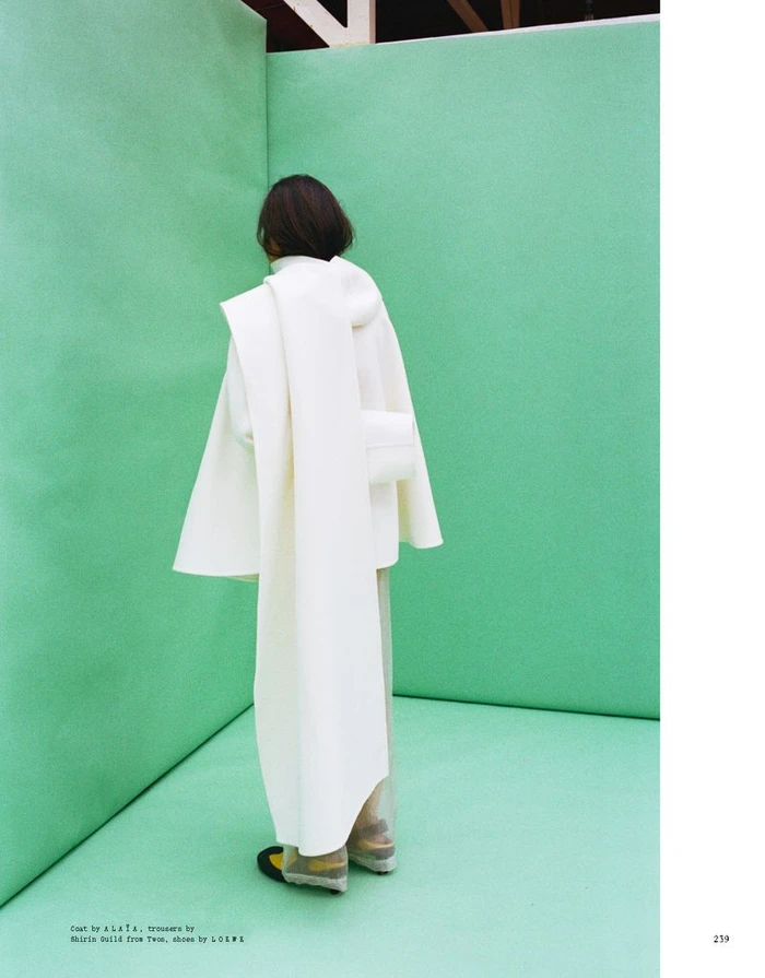Model seen from behind wearing an oversized white coat and scarf standing in a corner with mint green walls
