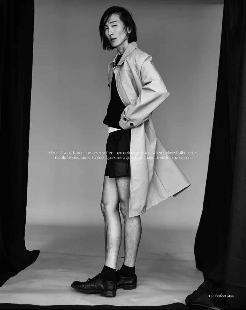Model Hyuk Kim in a beige trench coat and black shorts posing in a studio for The Perfect Man Magazine