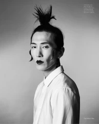 Monochrome portrait of model Hyuk Kim wearing dark lipstick and a white shirt with hair styled in a top knot against a grey background.