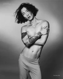 Monochrome portrait of model Hyuk Kim with tattooed arms wearing patterned trousers against a grey gradient background.