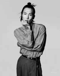 Model with hair in top knot wearing striped button-down shirt and trousers resting chin on hand against grey seamless background.