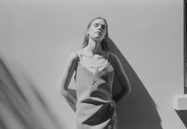Monochrome portrait of a model in a slip dress leaning against a wall outdoors with dappled sunlight.