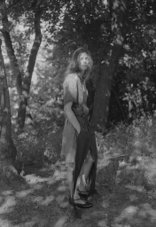 Model standing outdoors among trees wearing a short sleeve top and dark trousers with Mary Jane shoes.