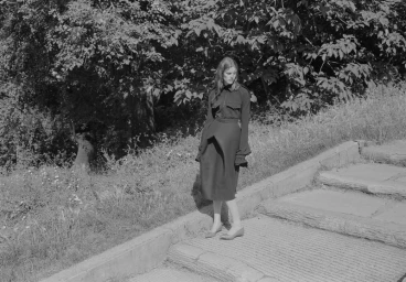 Model standing on stone steps near a wooded path wearing a dark long-sleeve dress and low heels