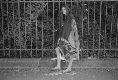 Model standing outdoors wearing a patterned poncho against a metal fence for Crosscurrent Another Way
