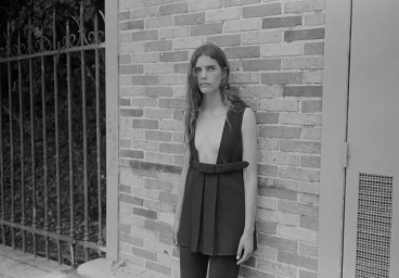 Model in a deep V-neck sleeveless black top leaning against a brick wall next to an ornate iron gate.