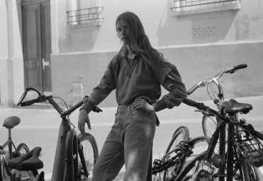 Model standing outdoors next to parked bicycles wearing a long sleeved top and trousers in a monochrome editorial photograph