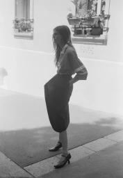 A woman standing outdoors in profile wearing a collared shirt, dark skirt, and kitten heels against a light building wall