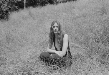 Monochrome image of a woman with long wavy hair crouching in a grassy meadow for the Crosscurrent Another Way project