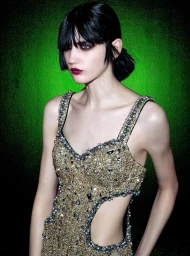 Model with dark hair and bangs wearing a heavily embellished gold crystal dress against a vibrant green textured background.