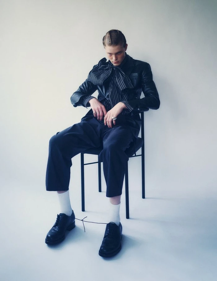Model seated on a black chair wearing a dark jacket, striped ascot, trousers, white socks, and black shoes against a plain white backdrop