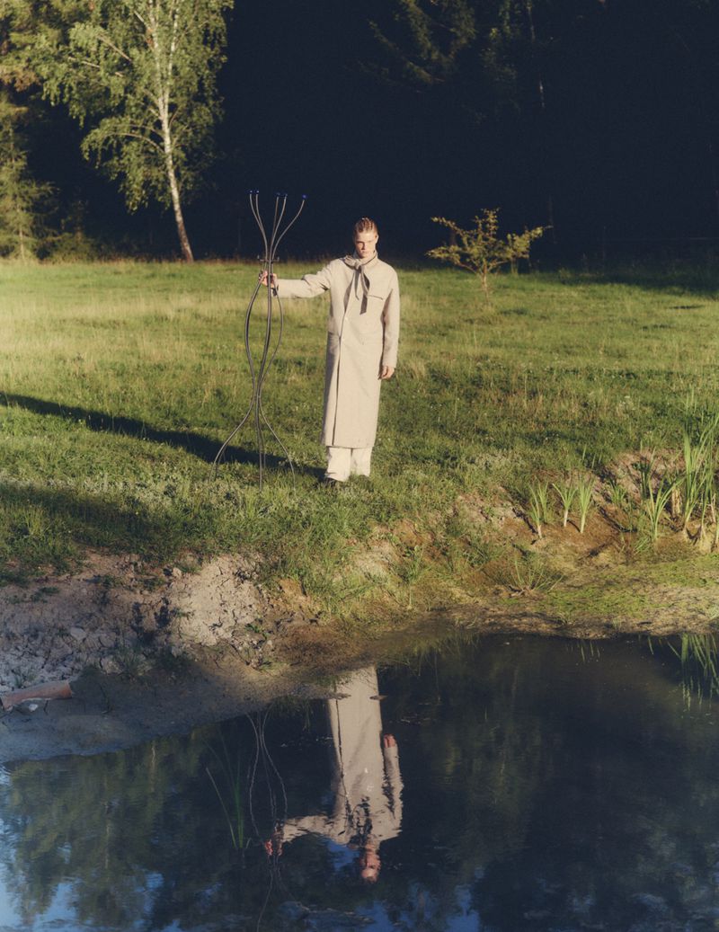 Model wearing a light beige coat holding a metal sculpture by a pond in a grassy meadow for Vogue Scandinavia