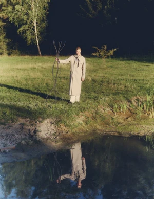 Model wearing a light beige coat holding a metal sculpture by a pond in a grassy meadow for Vogue Scandinavia
