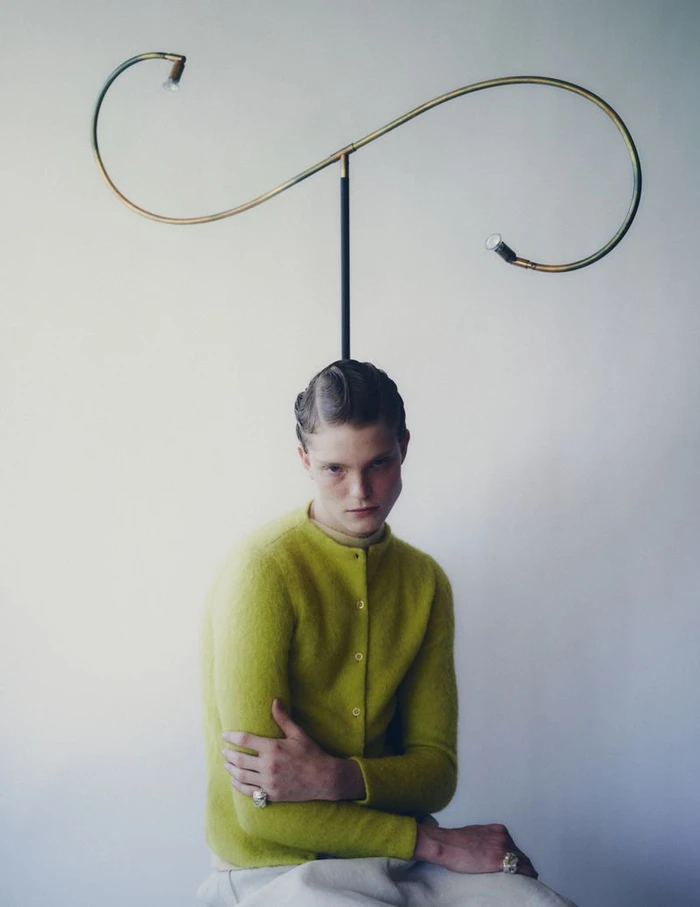 Model wearing a chartreuse green cardigan seated against a plain wall with a sculptural lamp framing his head.