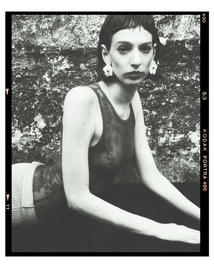 Monochrome fashion portrait of Brenda Pivatto wearing flower earrings and a patterned top against a textured wall.