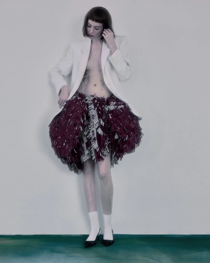 Model wearing an open white blazer and a textured burgundy skirt against a plain wall for Flanelle Magazine.