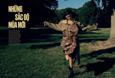A model wearing a brown coat and scarf running through a sunlit park with trees and green grass.