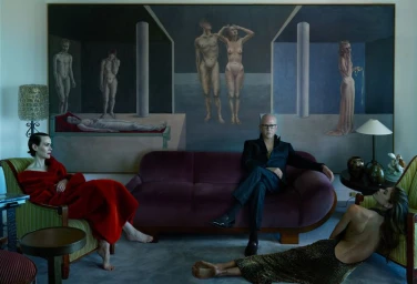 Ryan Murphy seated on a purple velvet sofa with Sarah Paulson in a red gown and another woman reclining on the floor.