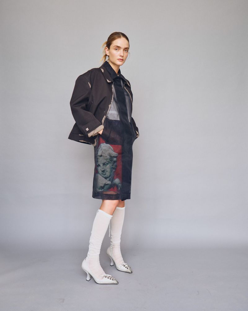 Model wearing dark structured jacket and printed dress with white knee-high boots against a grey seamless studio backdrop.