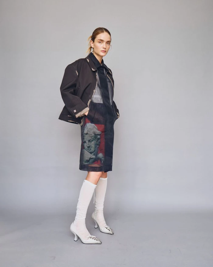 Model wearing dark structured jacket and printed dress with white knee-high boots against a grey seamless studio backdrop.
