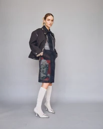 Model wearing dark structured jacket and printed dress with white knee-high boots against a grey seamless studio backdrop.